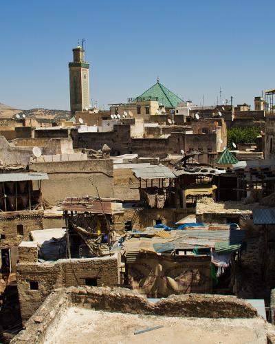 A high-angle view of a dense, historic urban district featuring traditional earthen buildings, a prominent square minaret, a green-tiled dome, and foreground rooftops with numerous satellite dishes and what appear to be dye vats.