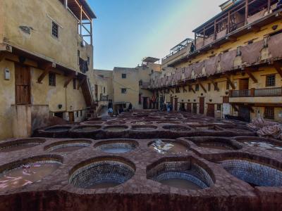 A high-angle view of a historic tannery courtyard featuring numerous hexagonal and circular stone vats filled with dyeing liquids, surrounded by multi-story buildings with wooden balconies.