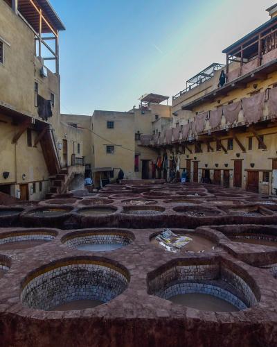 A high-angle view of a historic tannery courtyard featuring numerous hexagonal and circular stone vats filled with dyeing liquids, surrounded by multi-story buildings with wooden balconies.