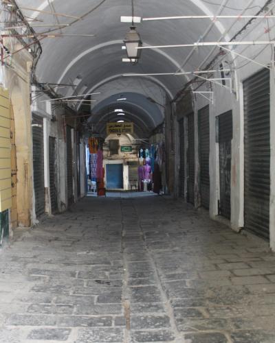 A long, vaulted stone passageway within a covered market, featuring closed metal shutters on the sides and a hanging sign in the distance.
