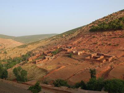 A cluster of traditional earthen houses built into a steep, reddish-brown hillside in a mountainous region, with terraced fields and scattered green vegetation under a clear sky.