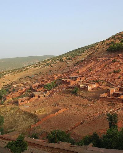 A cluster of traditional earthen houses built into a steep, reddish-brown hillside in a mountainous region, with terraced fields and scattered green vegetation under a clear sky.