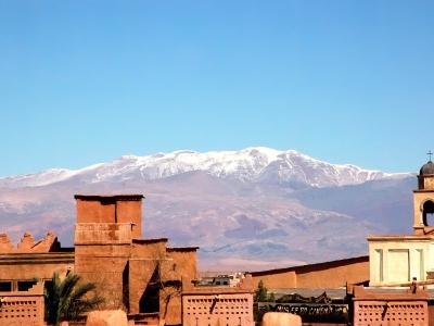 A view of traditional earthen architecture in the foreground with a snow-capped mountain range in the background, featuring a sign for "MUSEE DU CINEMA" on a building to the right.