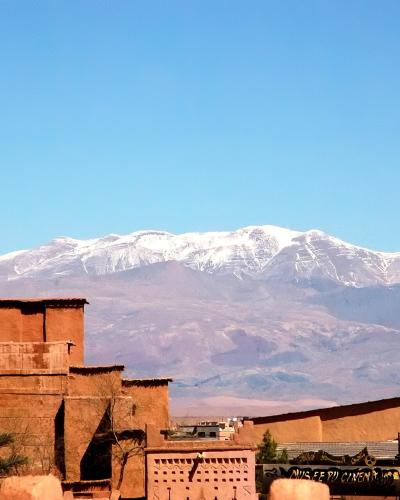 A view of traditional earthen architecture in the foreground with a snow-capped mountain range in the background, featuring a sign for "MUSEE DU CINEMA" on a building to the right.