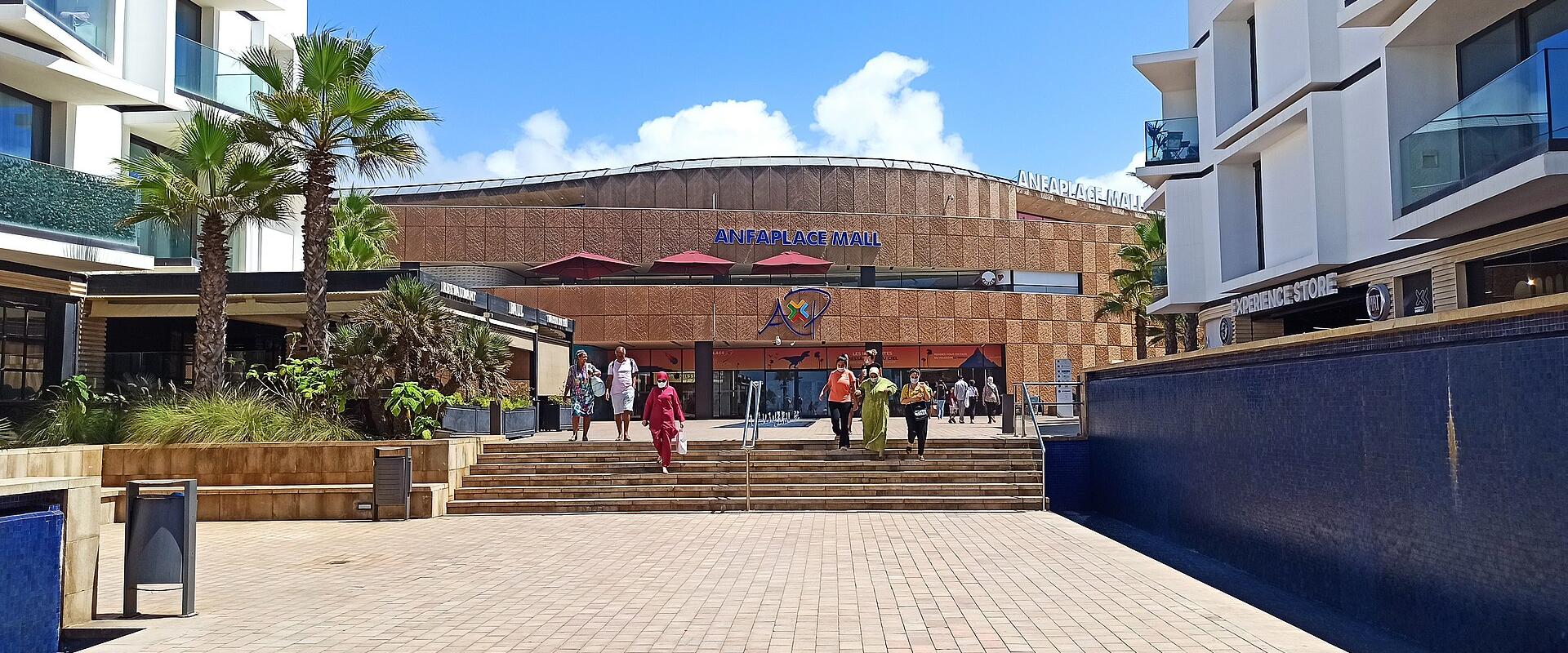 A wide-angle outdoor shot of the AnfaPlace Mall entrance in Casablanca, Morocco, showing its brown tiled facade, blue signage reading "ANFAPLACE MALL", palm trees, and shoppers on steps under a clear sky.