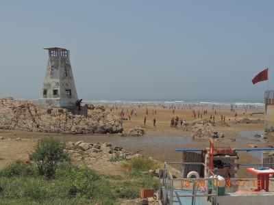 A sandy beach scene featuring a distinctive white, weathered concrete watchtower on a rocky outcrop to the left, a Moroccan flag flying on the right, and a crowded shoreline with the Atlantic Ocean in the background.