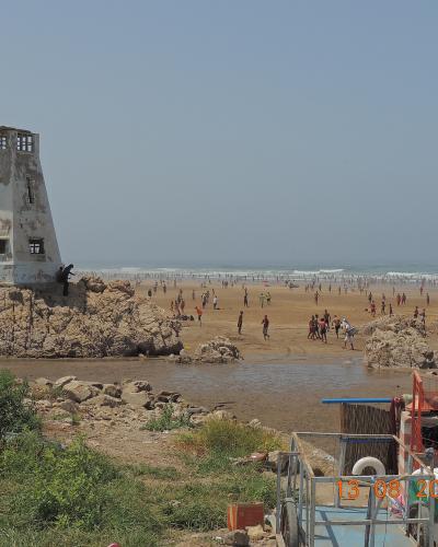 A sandy beach scene featuring a distinctive white, weathered concrete watchtower on a rocky outcrop to the left, a Moroccan flag flying on the right, and a crowded shoreline with the Atlantic Ocean in the background.