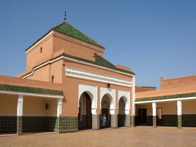 A courtyard view of a traditional Moroccan religious complex featuring terracotta walls, green-tiled hip roofs, white stucco arches, and intricate green and white zellige tilework on the lower walls and columns.