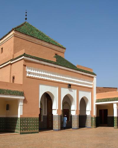 A courtyard view of a traditional Moroccan religious complex featuring terracotta walls, green-tiled hip roofs, white stucco arches, and intricate green and white zellige tilework on the lower walls and columns.