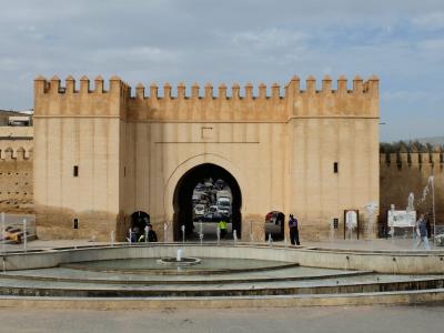 A large, beige stone gatehouse with crenellated battlements and a central archway, situated behind a tiered fountain in an open plaza.