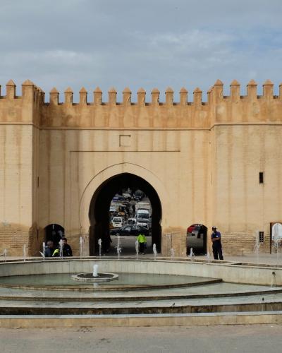 A large, beige stone gatehouse with crenellated battlements and a central archway, situated behind a tiered fountain in an open plaza.