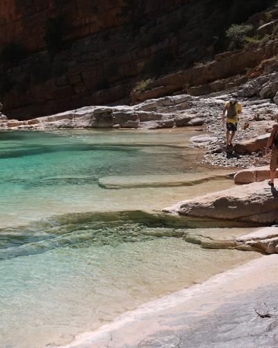 A group of tourists standing on rocky ledges beside a pool of clear, turquoise water within a narrow, sunlit canyon with layered sandstone walls.