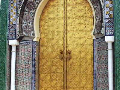 A close-up photograph of an ornate, arched doorway featuring large, embossed golden double doors set within a frame of intricate blue, white, and yellow zellige tilework and flanked by white marble columns.