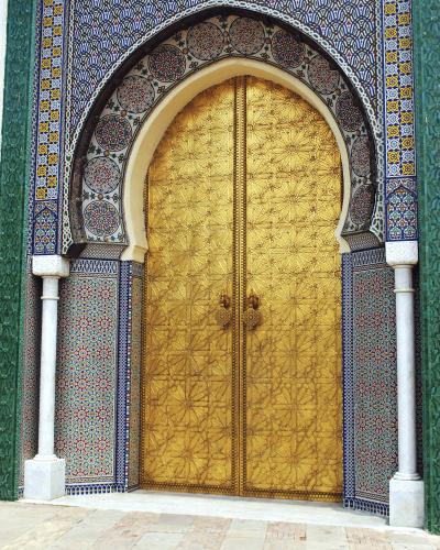 A close-up photograph of an ornate, arched doorway featuring large, embossed golden double doors set within a frame of intricate blue, white, and yellow zellige tilework and flanked by white marble columns.