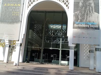 The image shows the main entrance of a modern white building with a large arched glass doorway, flanked by two vertical banners advertising art exhibitions, with Arabic and French text visible on the glass doors and banners.