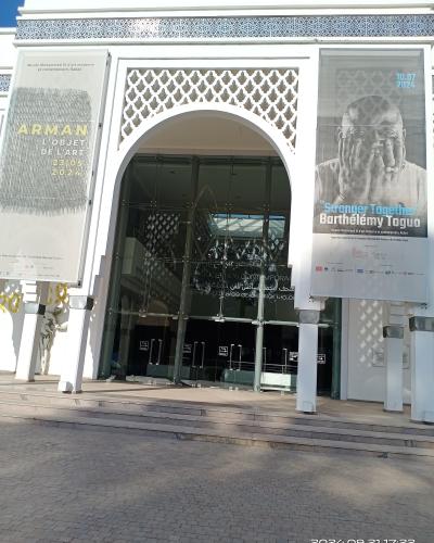 The image shows the main entrance of a modern white building with a large arched glass doorway, flanked by two vertical banners advertising art exhibitions, with Arabic and French text visible on the glass doors and banners.