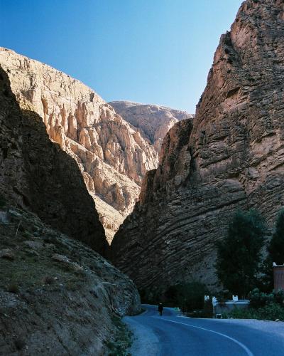 A paved road curves through a narrow, steep-sided canyon with layered sedimentary rock formations under a clear blue sky, with a lone figure walking on the road and some vegetation and structures visible on the right bank.