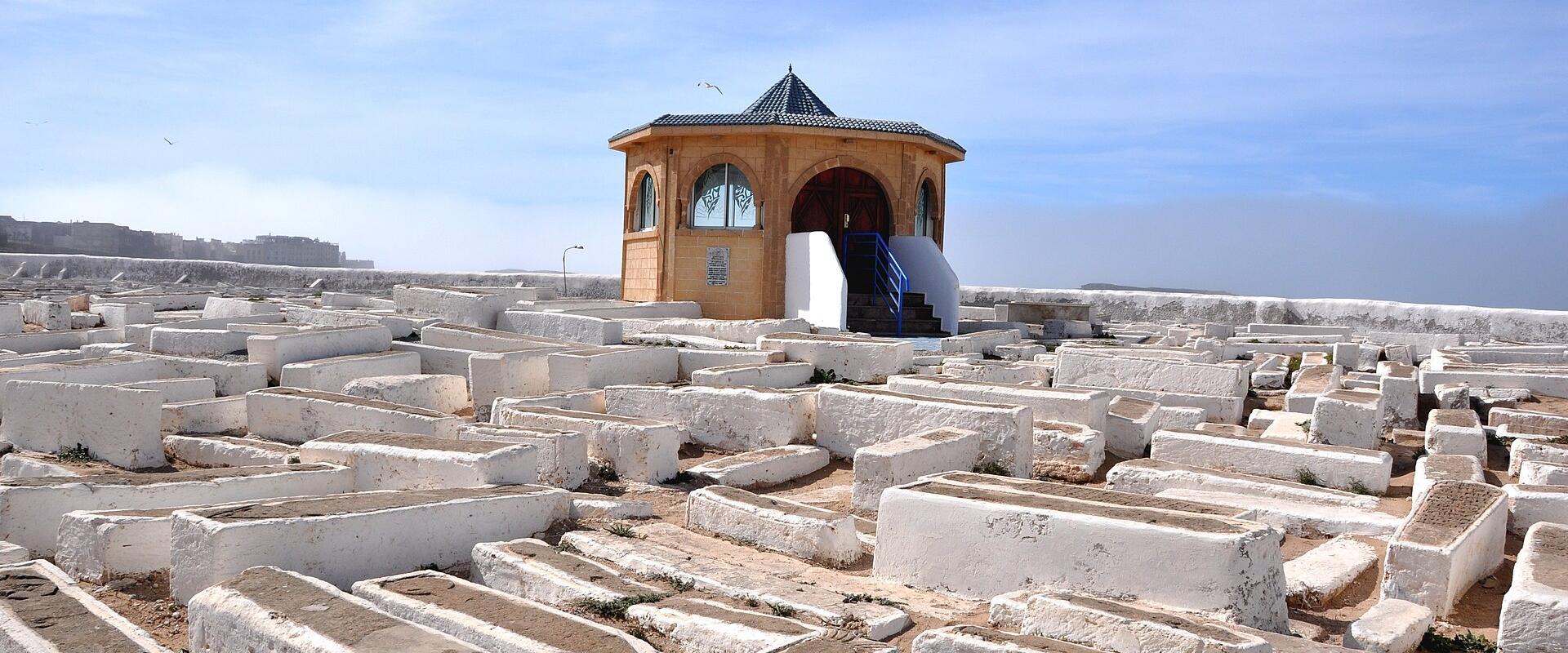 A cemetery with numerous white, rectangular tombs arranged in rows, centered around a small octagonal mausoleum with a blue-tiled roof and arched windows, set against a coastal backdrop with the ocean visible in the distance.