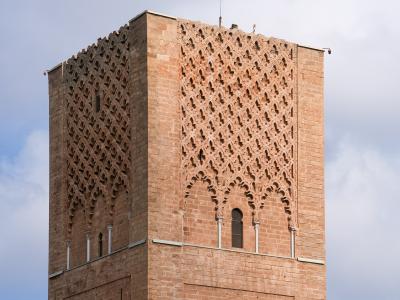 A low-angle shot of the upper section of a large, unfinished square minaret constructed from reddish-brown stone, featuring intricate geometric lattice patterns and horseshoe arches against a cloudy sky.