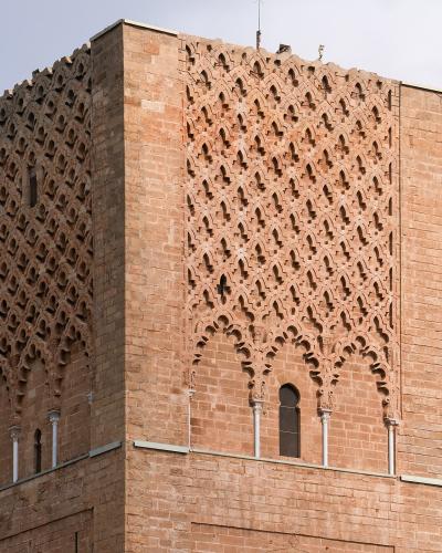 A low-angle shot of the upper section of a large, unfinished square minaret constructed from reddish-brown stone, featuring intricate geometric lattice patterns and horseshoe arches against a cloudy sky.
