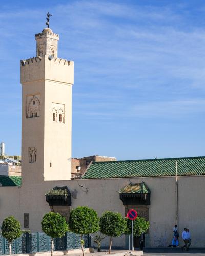 A tall, square minaret with crenellated battlements and arched windows stands next to a mosque building with a green-tiled roof, set against a blue sky with people walking on the street in the foreground.