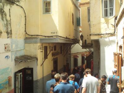 A narrow, sunlit alleyway in a historic medina with yellow and white stucco buildings, tourists walking away from the camera, and a sign reading "PUB ALLERAN GRANDSUDO" on a wall.