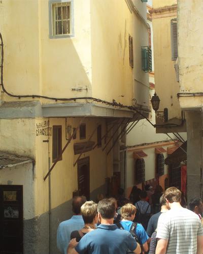 A narrow, sunlit alleyway in a historic medina with yellow and white stucco buildings, tourists walking away from the camera, and a sign reading "PUB ALLERAN GRANDSUDO" on a wall.