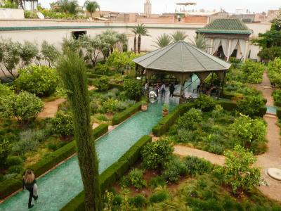 A high-angle view of a lush, manicured garden featuring a long rectangular pool with green tiles, a central gazebo, and traditional Moroccan architecture in the background.