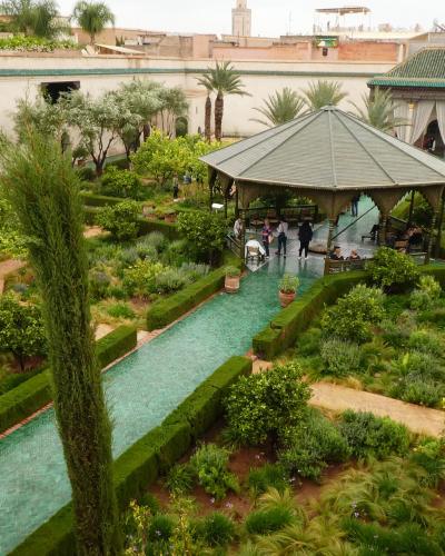 A high-angle view of a lush, manicured garden featuring a long rectangular pool with green tiles, a central gazebo, and traditional Moroccan architecture in the background.