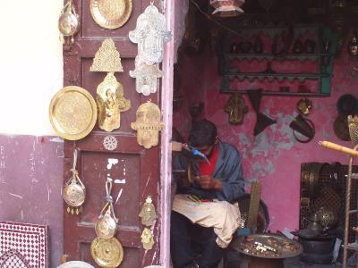 A craftsman sits in a doorway working on metal goods, surrounded by hanging brass plates, lanterns, and silver teapots displayed on the street.