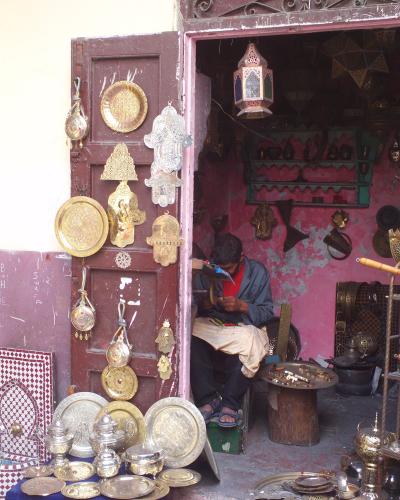 A craftsman sits in a doorway working on metal goods, surrounded by hanging brass plates, lanterns, and silver teapots displayed on the street.