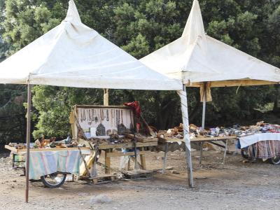Two white canvas tents set up outdoors in a wooded area, displaying various handicrafts like jewelry and wooden items on tables and a cart.