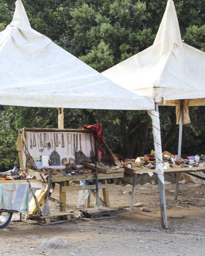 Two white canvas tents set up outdoors in a wooded area, displaying various handicrafts like jewelry and wooden items on tables and a cart.