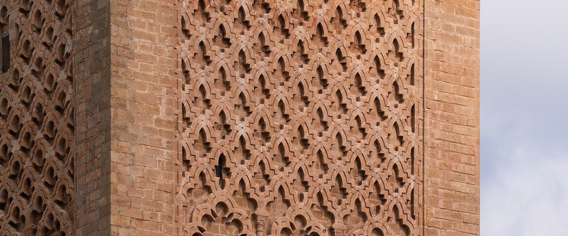 A close-up view of the upper corner of a large, unfinished sandstone minaret featuring intricate geometric brickwork and horseshoe arch motifs against a cloudy sky.