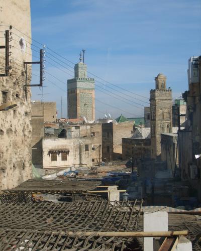 A view of rooftops and narrow alleys in a historic North African city, featuring two prominent minarets—one with green zellige tilework and the other with stone masonry—under a clear blue sky.
