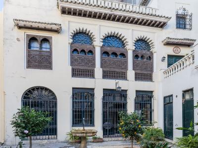 A courtyard view of a white-washed building featuring Moroccan-style wooden mashrabiya screens, arched windows with iron grilles, and a central stone fountain surrounded by potted citrus trees.