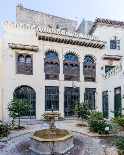 A courtyard view of a white-washed building featuring Moroccan-style wooden mashrabiya screens, arched windows with iron grilles, and a central stone fountain surrounded by potted citrus trees.