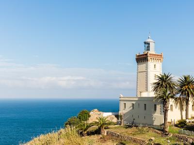 A tall, cream-colored lighthouse with a distinctive reddish-brown decorative band near the lantern room, situated on a grassy cliff overlooking the ocean, with palm trees in the foreground.
