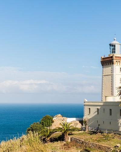 A tall, cream-colored lighthouse with a distinctive reddish-brown decorative band near the lantern room, situated on a grassy cliff overlooking the ocean, with palm trees in the foreground.