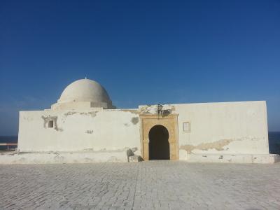 A white-washed, single-story religious building with a central dome and a horseshoe arch entrance, situated on a paved terrace overlooking the ocean under a clear blue sky.