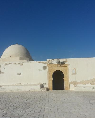 A white-washed, single-story religious building with a central dome and a horseshoe arch entrance, situated on a paved terrace overlooking the ocean under a clear blue sky.