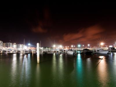 A night-time long-exposure photograph of a modern marina with white yachts docked at piers, illuminated by bright artificial lights that create starburst effects and reflections on the calm water, with multi-story buildings visible on the left shore.