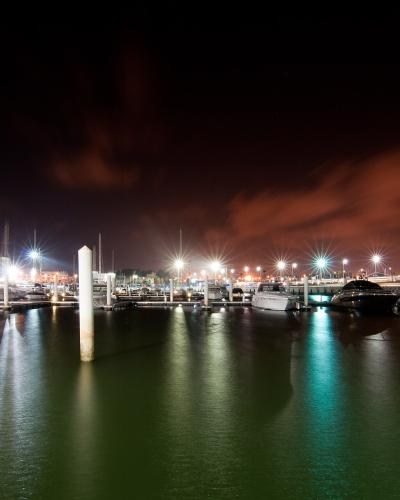 A night-time long-exposure photograph of a modern marina with white yachts docked at piers, illuminated by bright artificial lights that create starburst effects and reflections on the calm water, with multi-story buildings visible on the left shore.