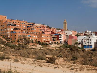 A hillside view of a densely built residential area with terracotta-colored apartment buildings and a mosque minaret, set against a clear blue sky with dry scrubland in the foreground.