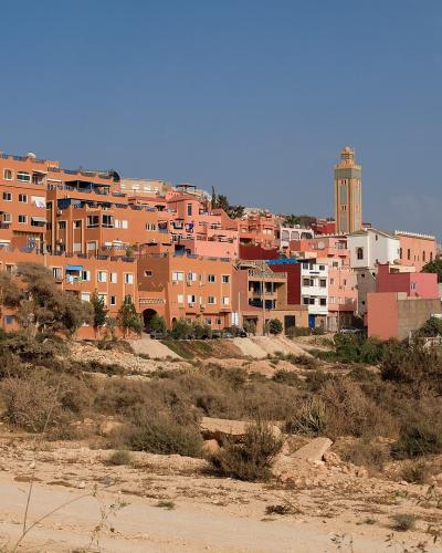 A hillside view of a densely built residential area with terracotta-colored apartment buildings and a mosque minaret, set against a clear blue sky with dry scrubland in the foreground.