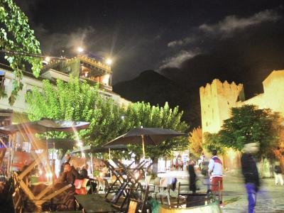 A night scene of a paved public square with outdoor cafe seating, trees, and a prominent stone fortress tower on the right side.