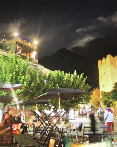 A night scene of a paved public square with outdoor cafe seating, trees, and a prominent stone fortress tower on the right side.