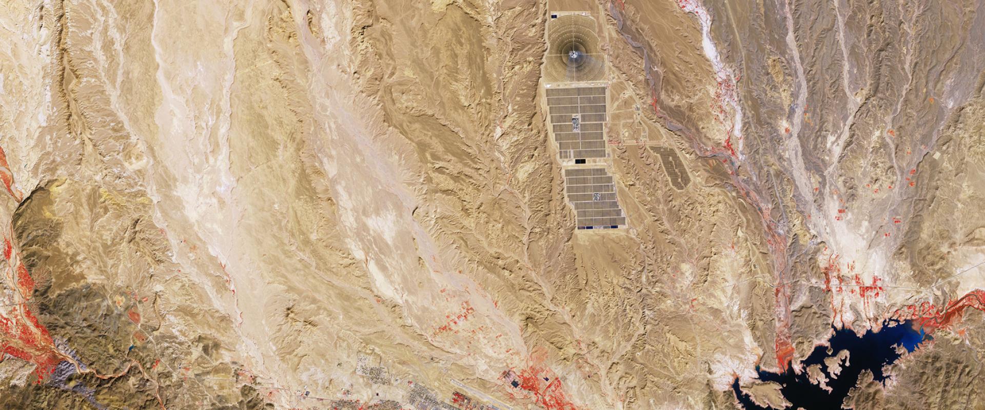 A satellite/aerial view of a desert landscape featuring a large solar power plant (Concentrated Solar Power with a central tower and heliostat field), a reservoir/lake, and a town, with false-color red highlighting vegetation or water features.