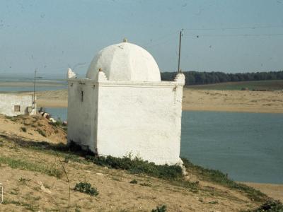 A white, domed religious structure (likely a zawiya or small mosque) situated on a sandy bank next to a body of water, with a line of trees in the background.
