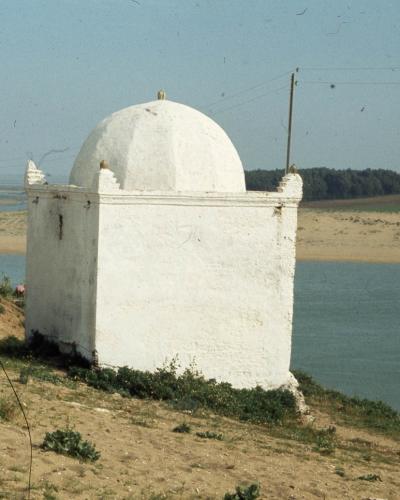 A white, domed religious structure (likely a zawiya or small mosque) situated on a sandy bank next to a body of water, with a line of trees in the background.